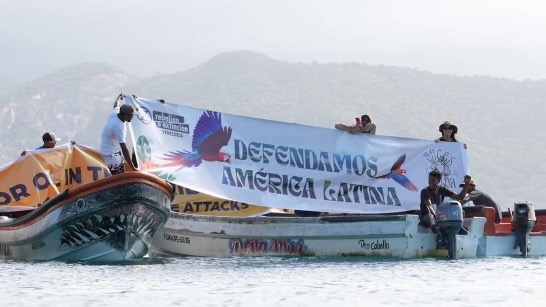 Una fotografia che riprende evidentemente le stesse barche e persone dellafoto precedente, ma da un'angolazione diversa e più ravvicinata. Le personereggono uno striscione bianco con la scritta "Defendamos AmericaLatina".