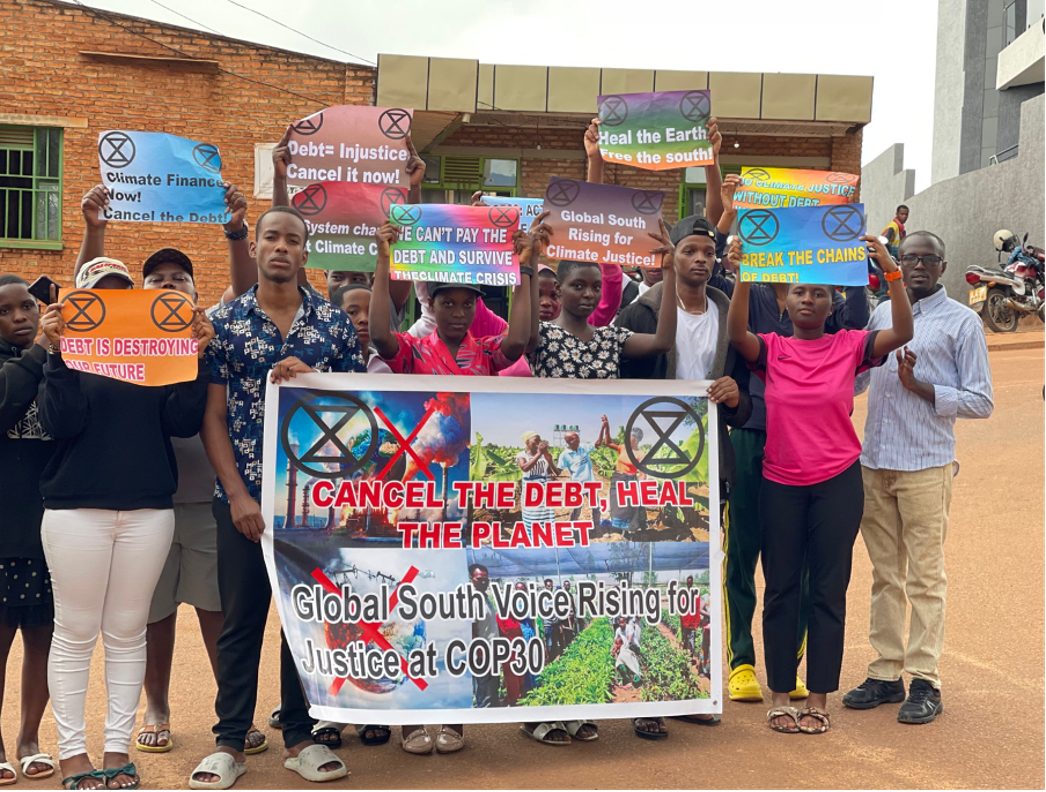 Uma foto de um grupo de pessoas em frente a um edifício de tijolos queseguram cartazes com símbolos do movimento XR e slogans relacionados. Ocartaz maior diz "cancel the debt heal theplanet".
