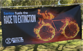 Fotografia de um banner que diz "Santos fuels the race to extinction" como logotipo da XR e uma bicicleta em chamas.